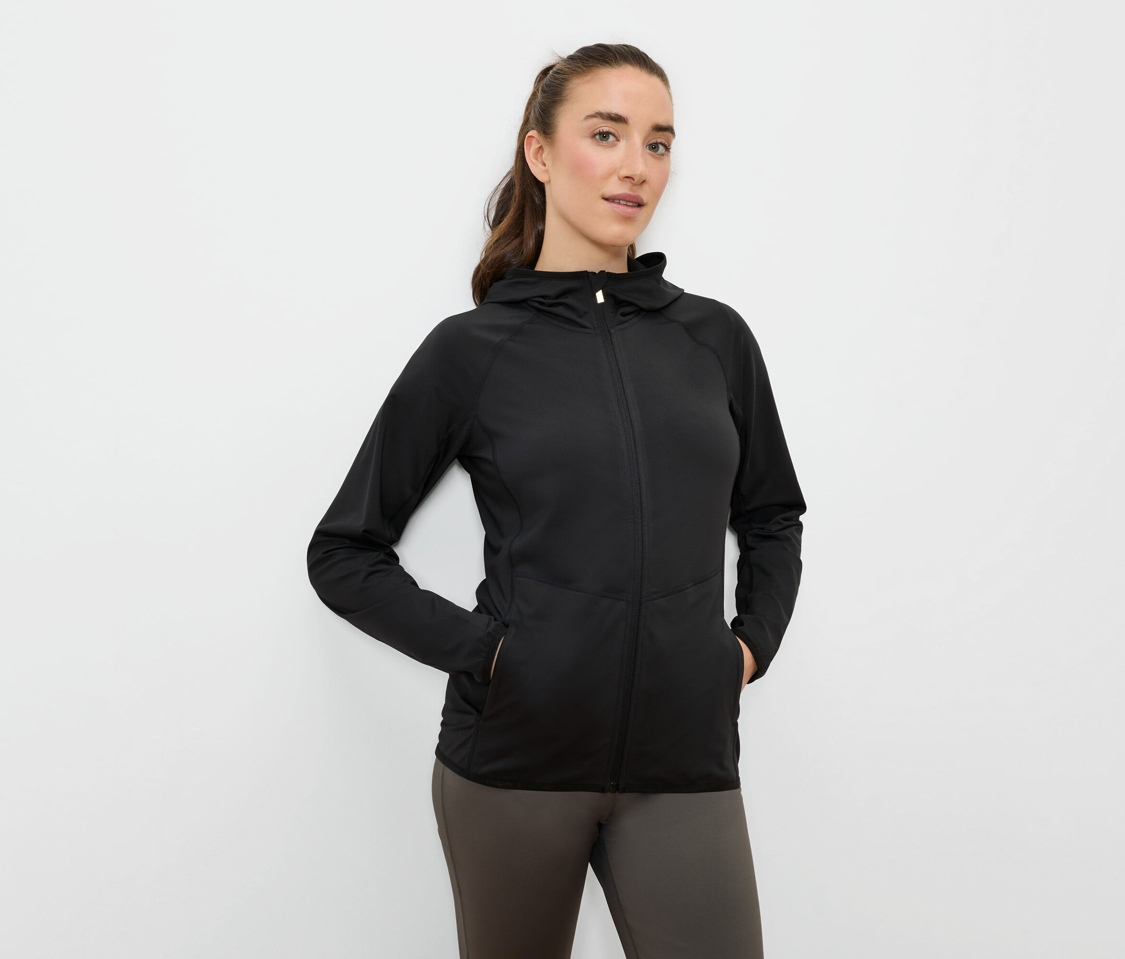 Kobieta trzyma ręce w kieszeniach czarnej sportowej kurtki z kapturem zapinanej na zamek.