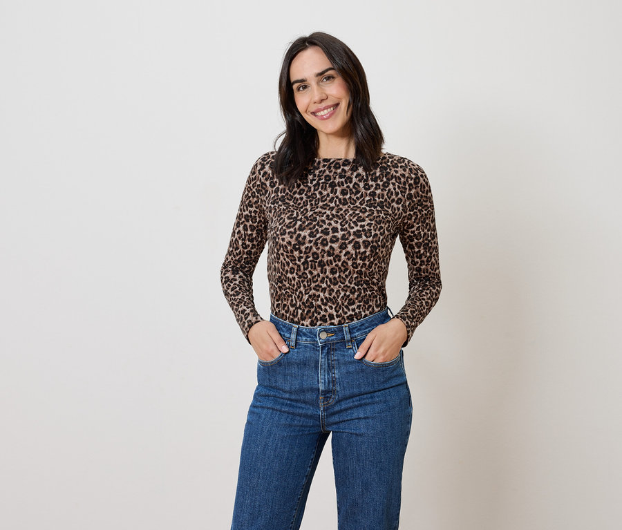 Kobieta o brązowych włosach pozuje w bluzce we wzór zwierzęcy i niebieskich dżinsach.