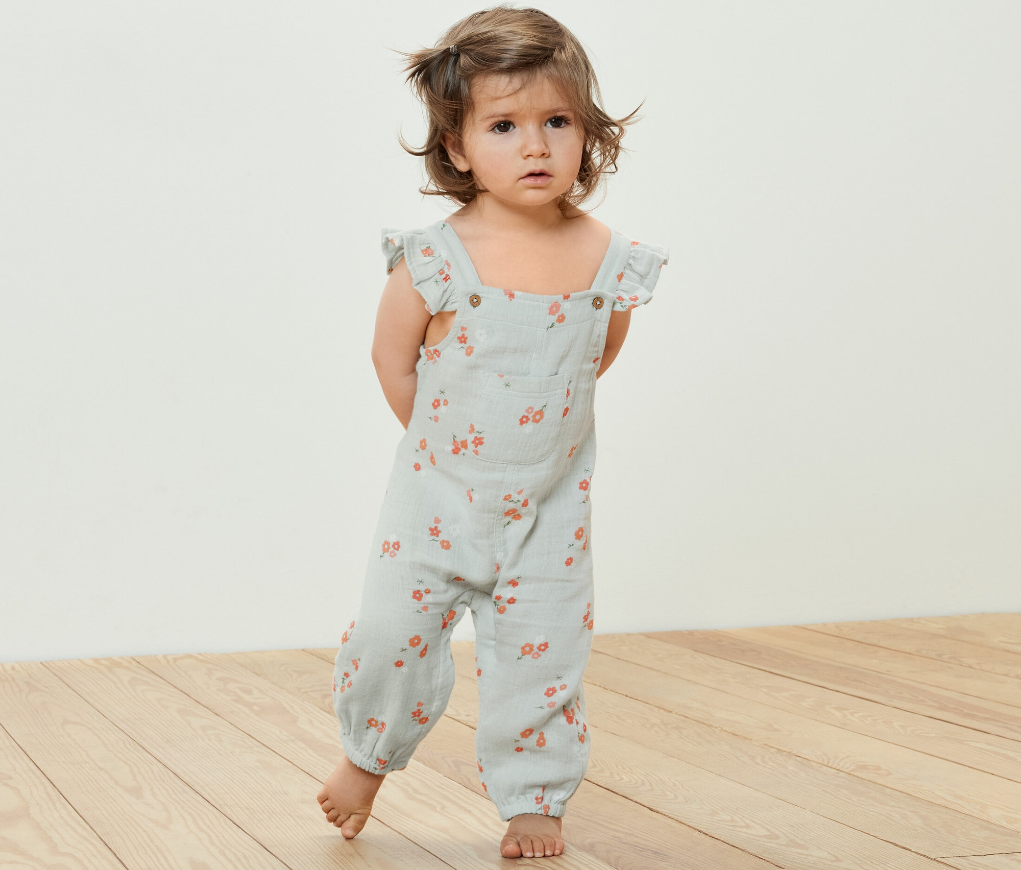 Małe dziecko stoi na drewnianej podłodze i ma na sobie muślinowy kombinezon w kwiatowy wzór.