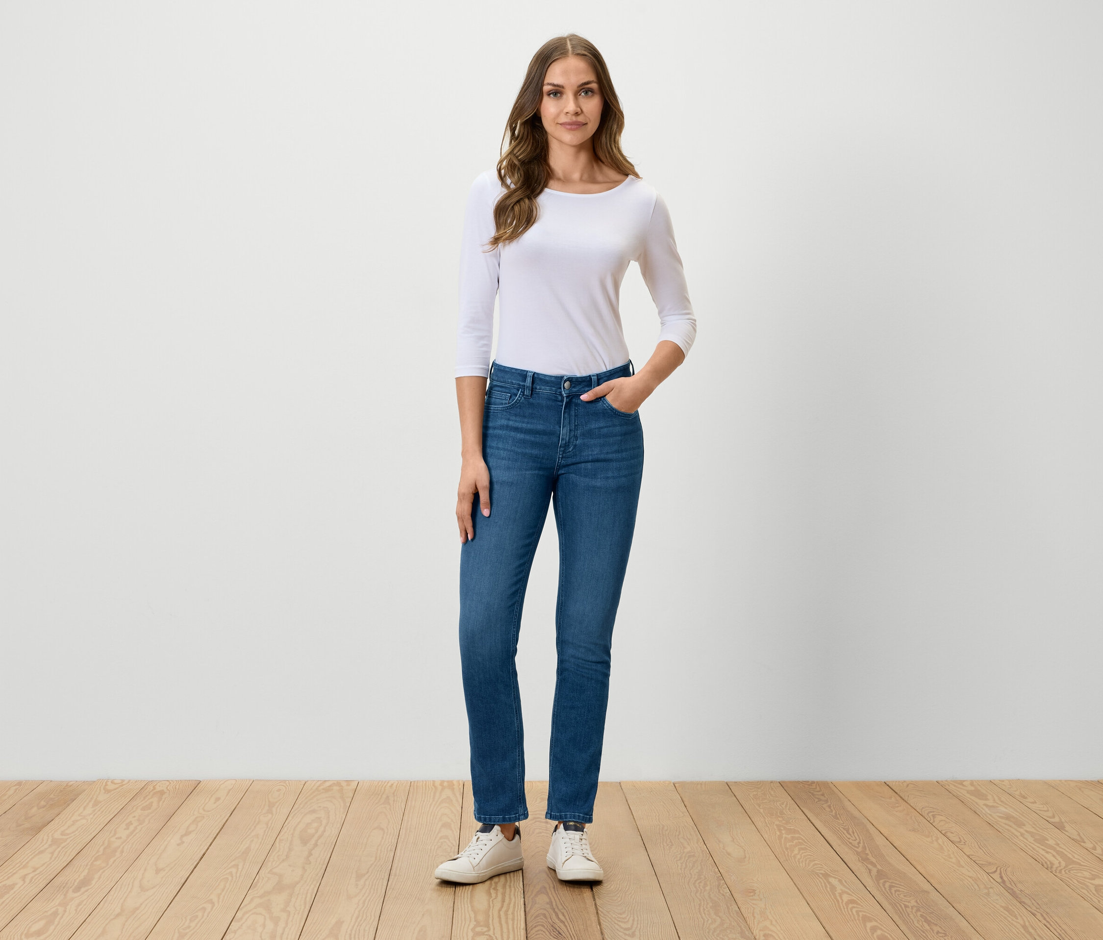 Kobieta stoi i pozuje w niebieskich dżinsach, białej koszulce i białych tenisówkach.