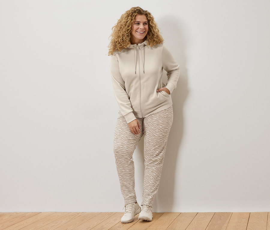 Kobieta z kręconymi włosami w beżowej bluzie z kapturem i spodniach w zebrę opiera się o białą ścianę.