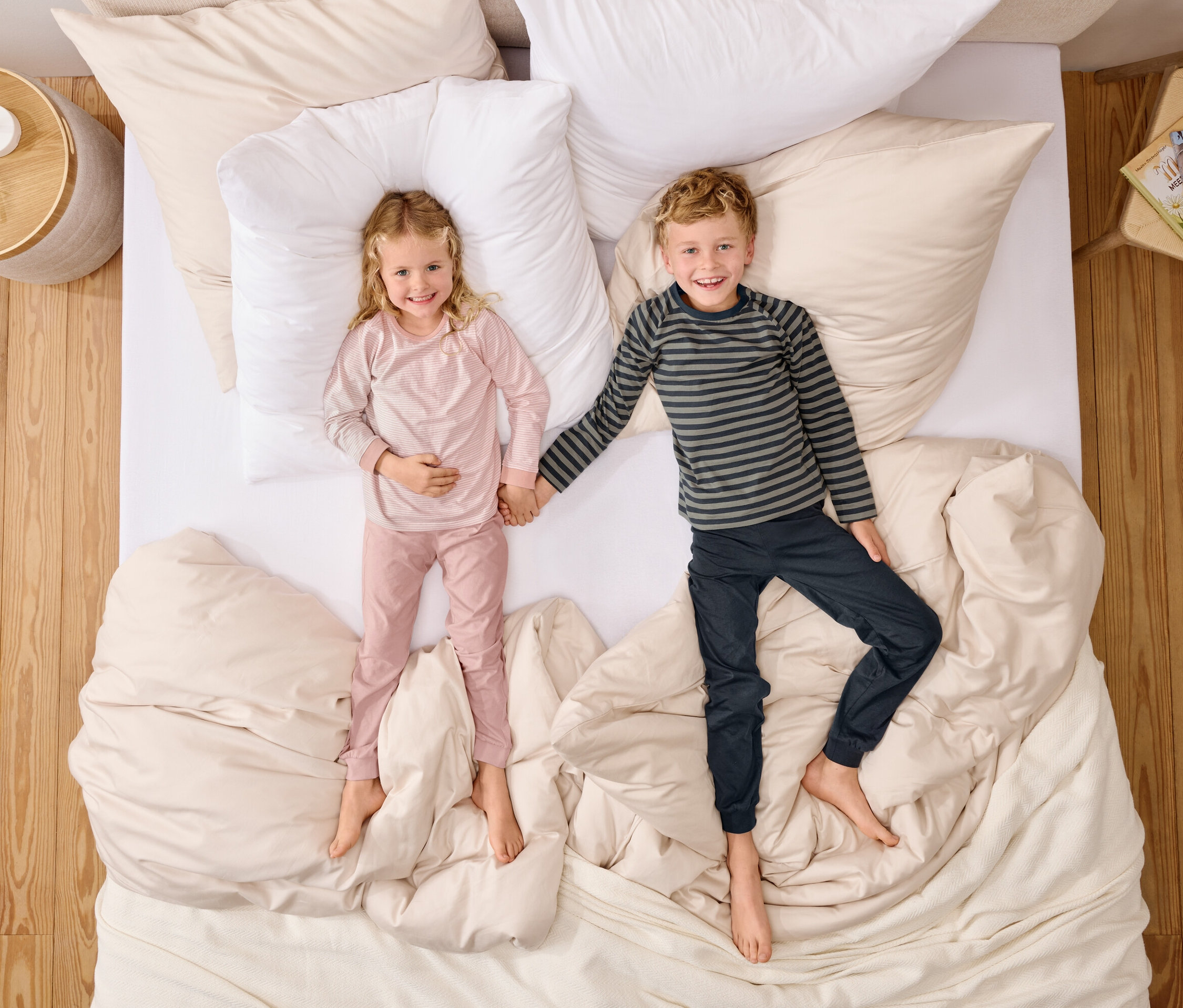 Widok z góry na dziewczynkę w różowej piżamie i chłopca w koszulce w paski, trzymających się za ręce na łóżku z białymi poduszkami.