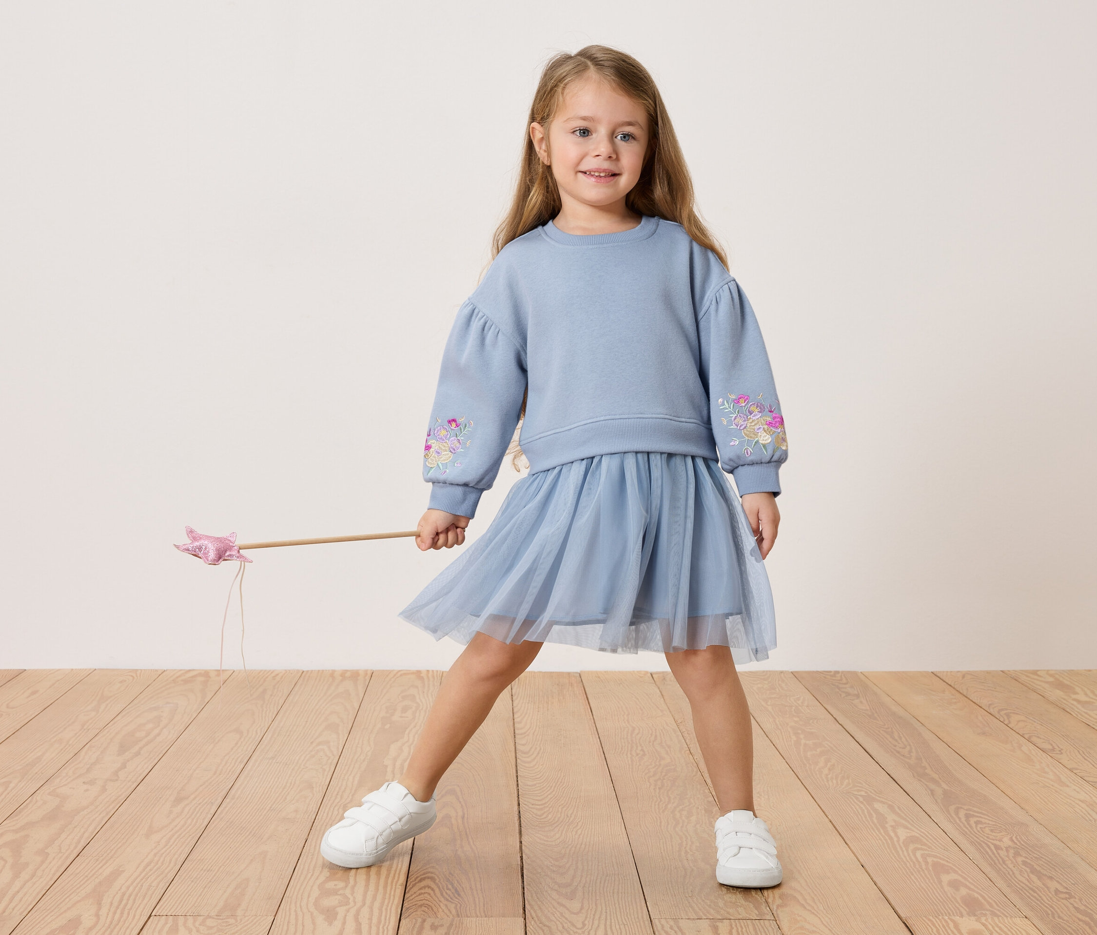 Dziewczynka w niebieskiej bluzie z kwiatowym haftem i tiulowej spódnicy, trzyma różdżkę.