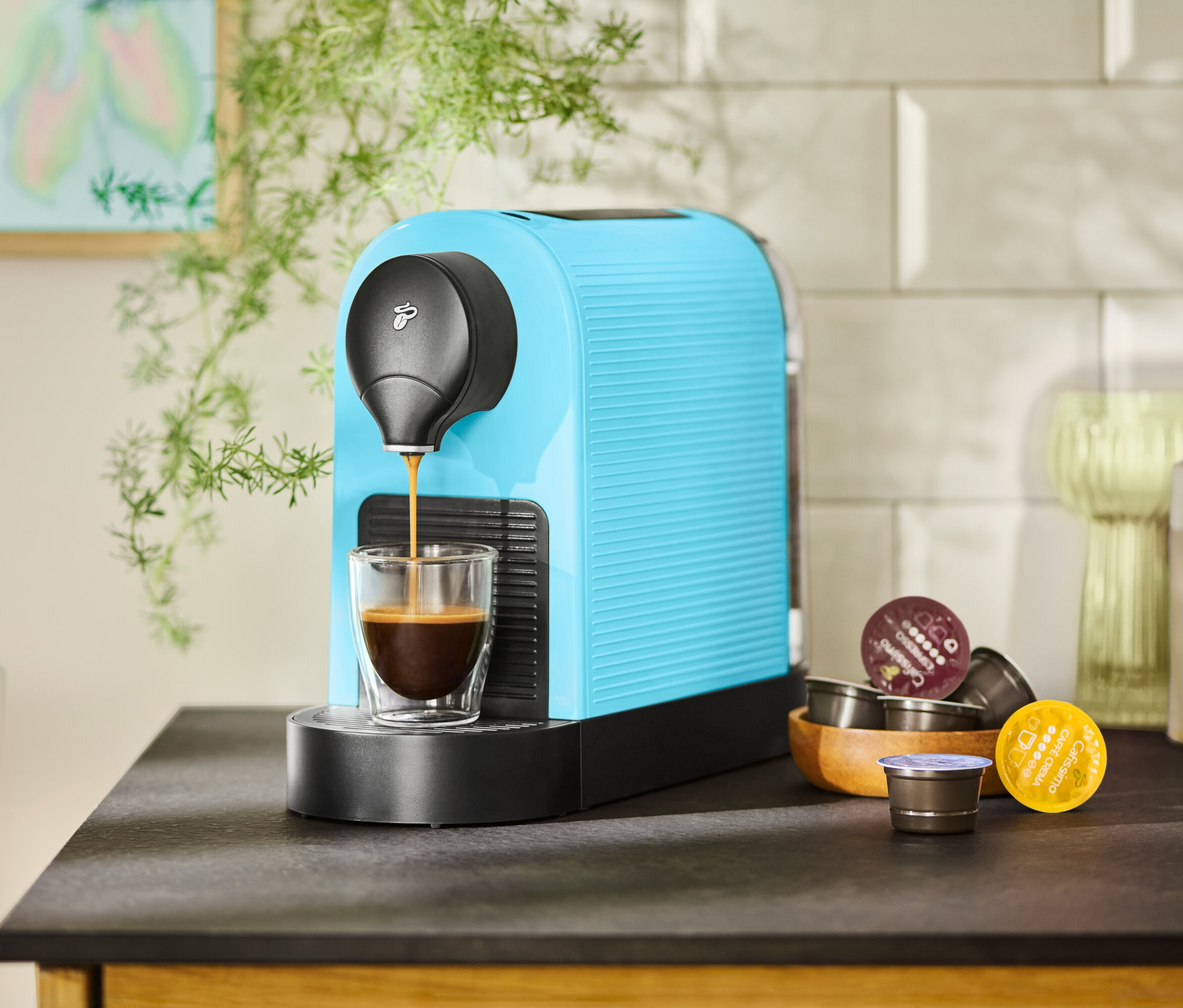 Turkusowy ekspres do kawy napełnia filiżankę kawą. Obok w drewnianej misce leżą kapsułki z kawą.