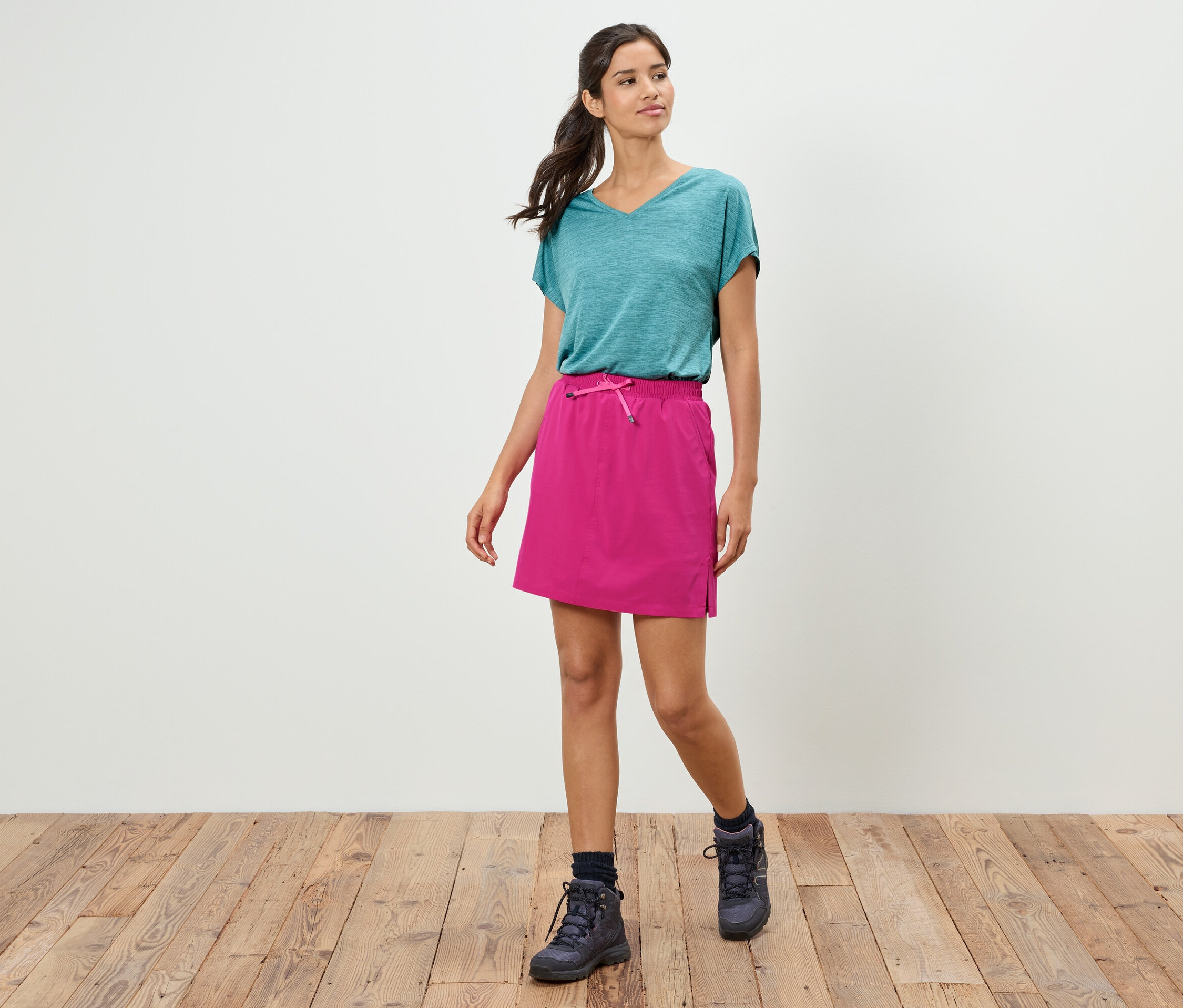 Kobieta idzie w turkusowej koszulce, różowej spódnicy i butach trekkingowych na drewnianej podłodze na tle białej ściany.