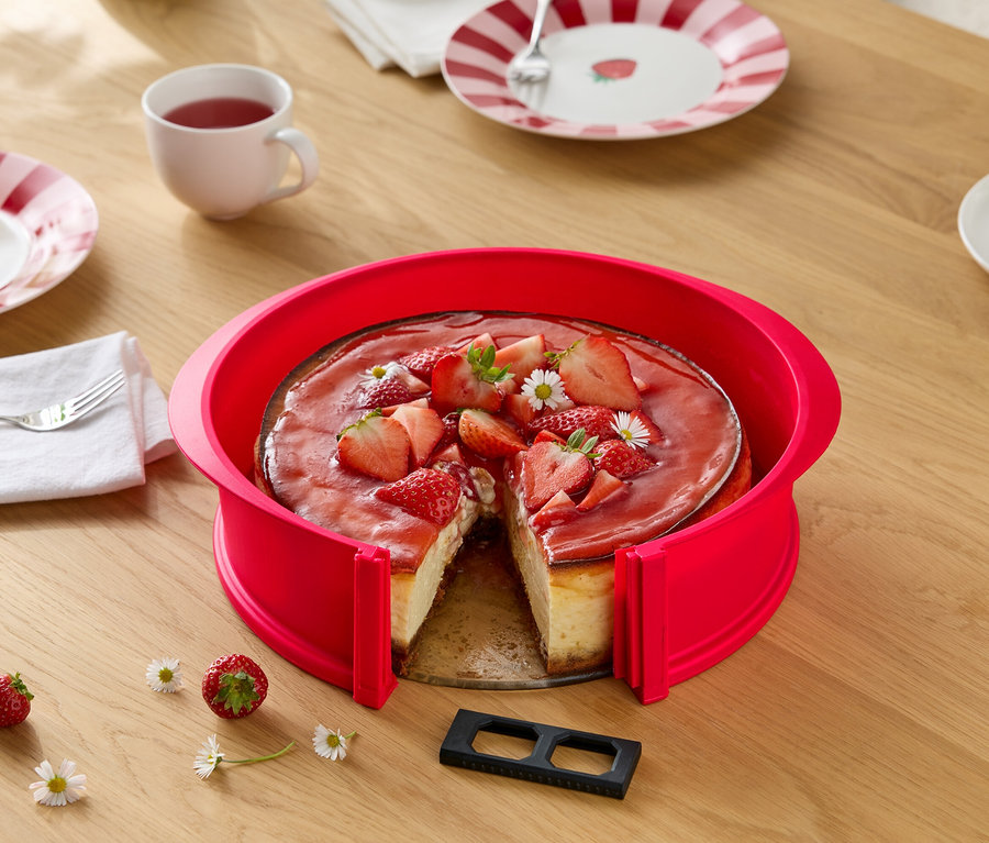 Krojony sernik truskawkowy w czerwonej formie na drewnianym stole z stokrotkami, nakryciem i napojami.