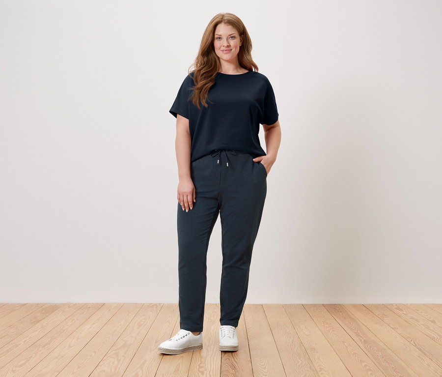 Kobieta stoi na tle ściany, ubrana w granatowy T-shirt, granatowe spodnie i białe buty.