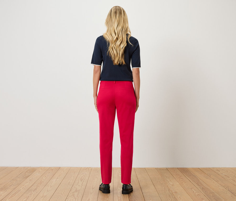 Kobieta o blond, falowanych włosach stoi tyłem do kamery, ubrana w granatową bluzkę i czerwone spodnie.