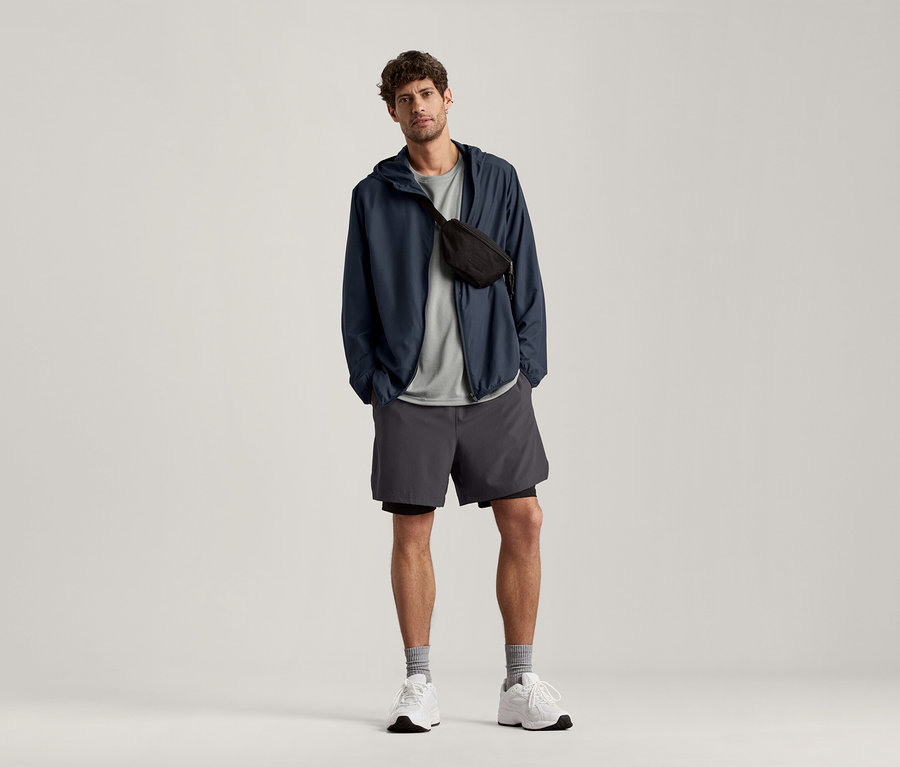 Mężczyzna stoi i ma na sobie jasno szarą koszulkę sportową, granatową kurtkę i szare szorty.
