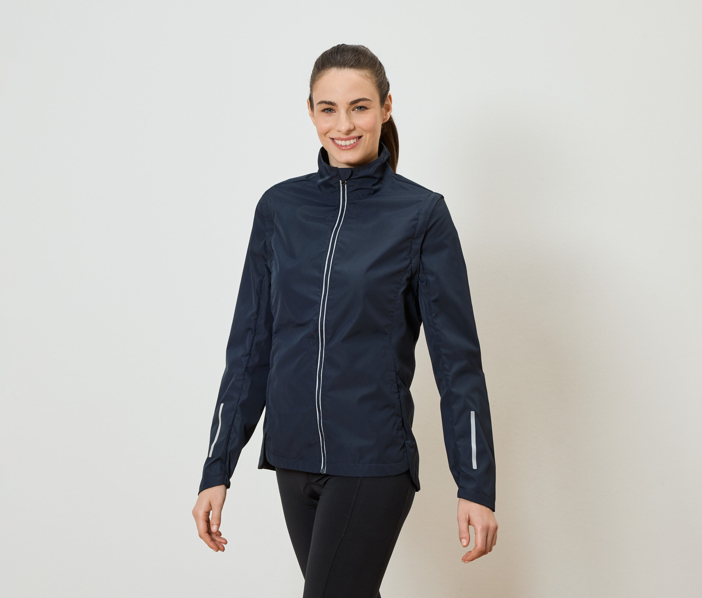 Kobieta z kucykiem ma na sobie granatową kurtkę zapinaną na zamek i czarne legginsy.