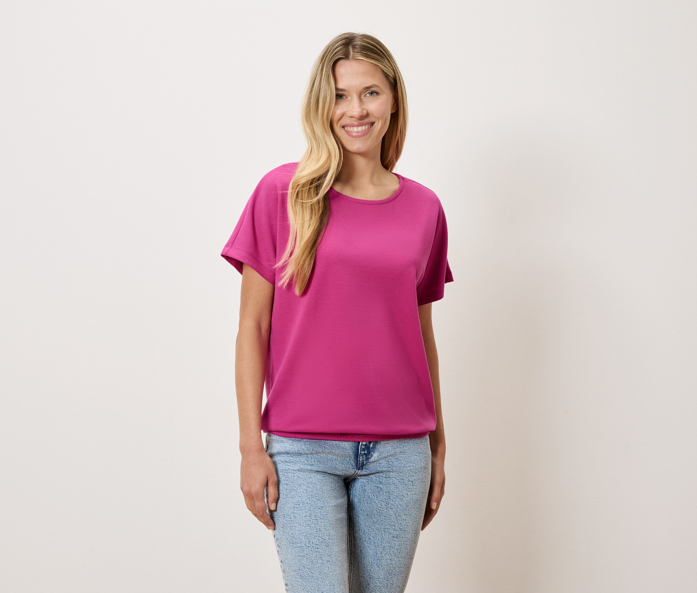 Kobieta o blond włosach ma na sobie różowy T-shirt i niebieskie dżinsy.