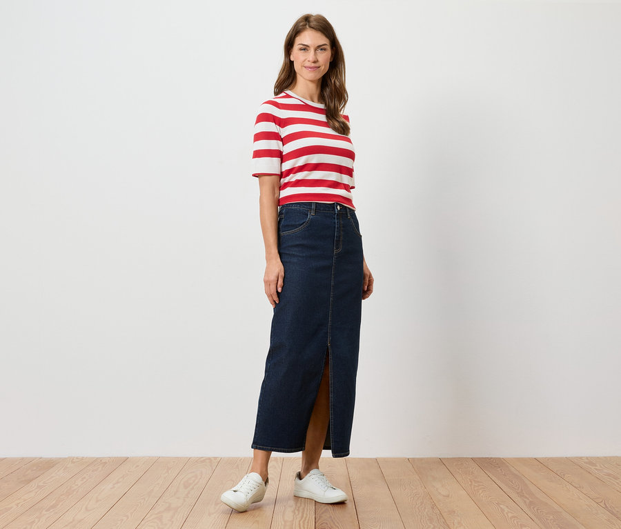 Kobieta stoi, ubrana w czerwono-białą koszulkę w paski, długą dżinsową spódnicę z rozcięciem i białe tenisówki.