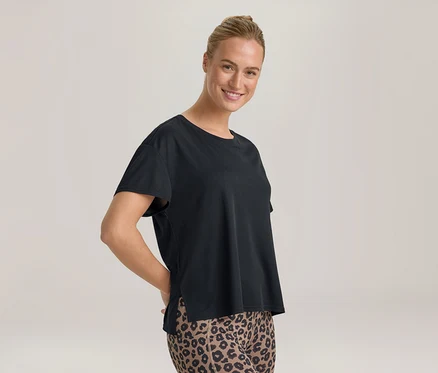 Uśmiechnięta kobieta z upiętymi włosami, ubrana w czarny T-shirt i legginsy w panterkę, z rękami za plecami.
