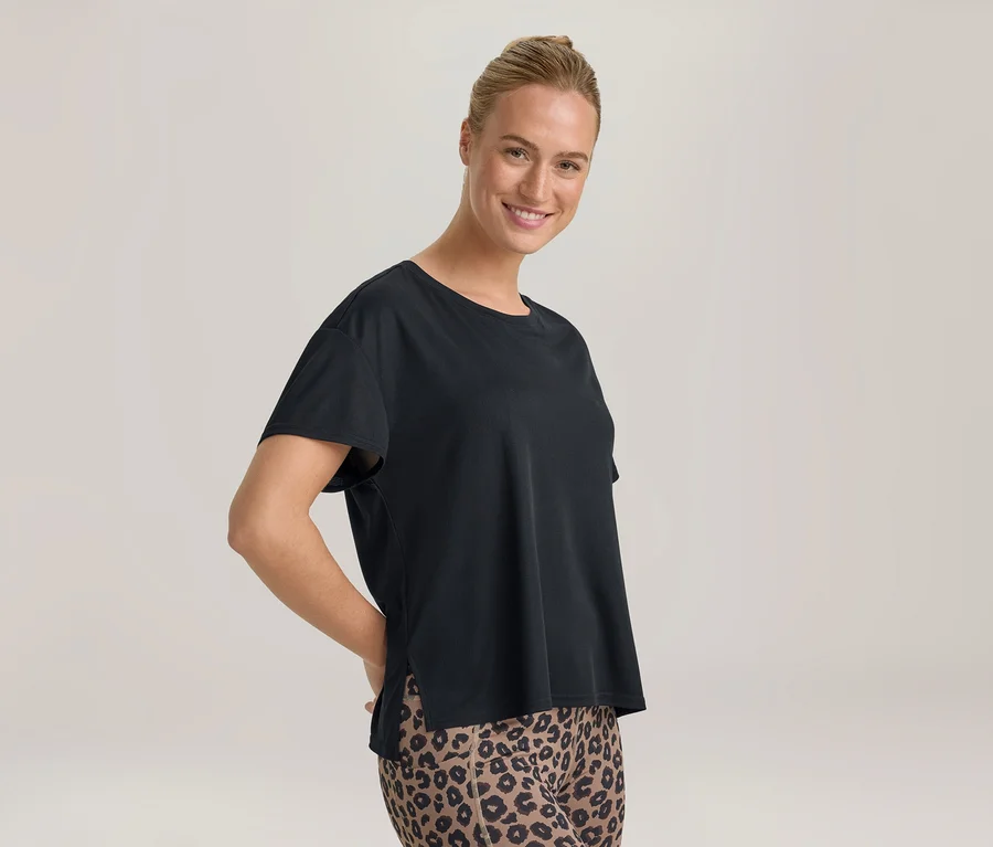 Uśmiechnięta kobieta z upiętymi włosami, ubrana w czarny T-shirt i legginsy w panterkę, z rękami za plecami.