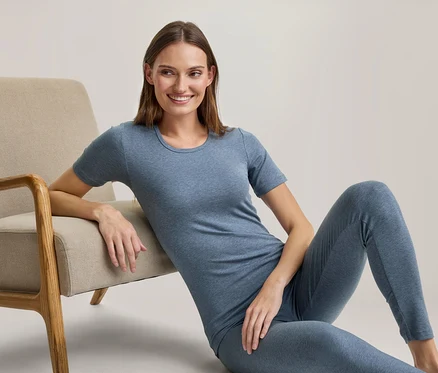 Uśmiechnięta kobieta siedzi na podłodze, opierając się o beżowy fotel, ubrana w niebieski T-shirt i pasujące legginsy.