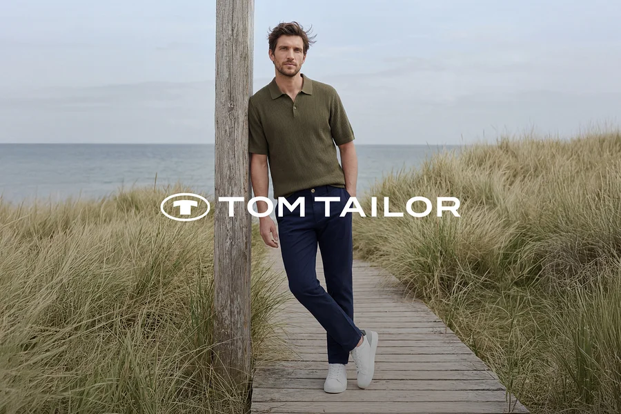 Mężczyzna w zielonej koszulce polo, granatowych spodniach i białych sneakersach opiera się o drewniany słup na pomoście przy morzu, otoczony wysoką trawą. Logo TOM TAILOR.