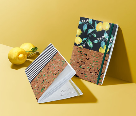 Dwa korkowe notatniki leżą obok dwóch cytryn na żółtej powierzchni.