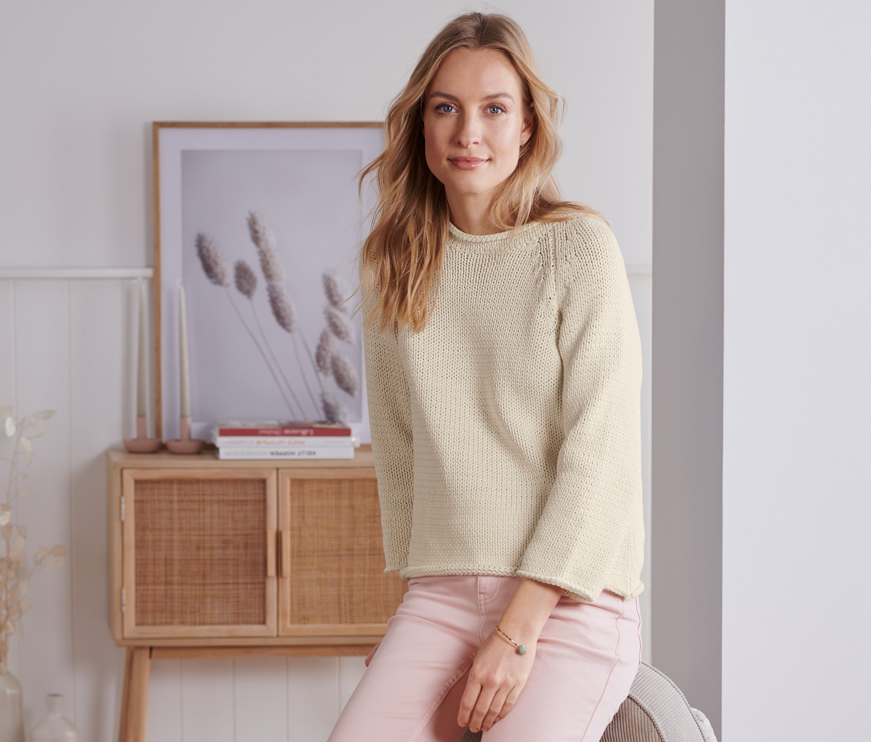 Kobieta siedzi na sofie i ma na sobie kremowy sweter o grubym splocie z przędzy tasiemkowej.