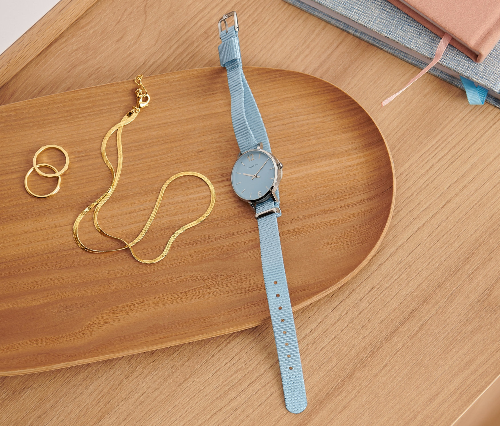 Niebieski zegarek kwarcowy ze stali nierdzewnej 316L leży na drewnianej tacy obok złotego naszyjnika i pierścionków.