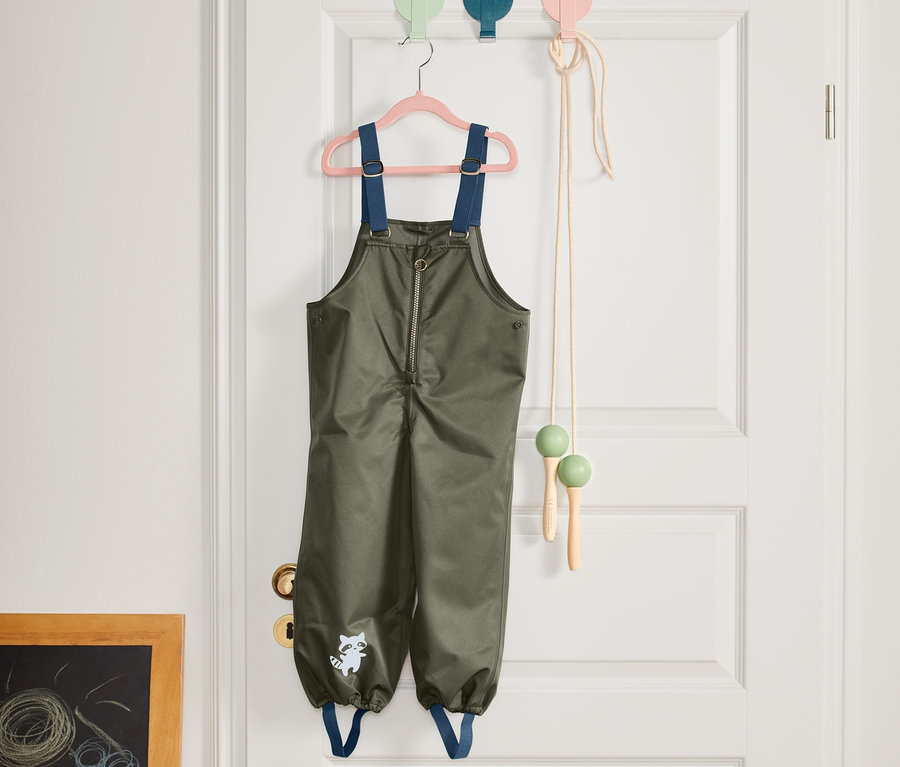 Zielone spodnie przeciwdeszczowe wiszą na różowym wieszaku na białych drzwiach.