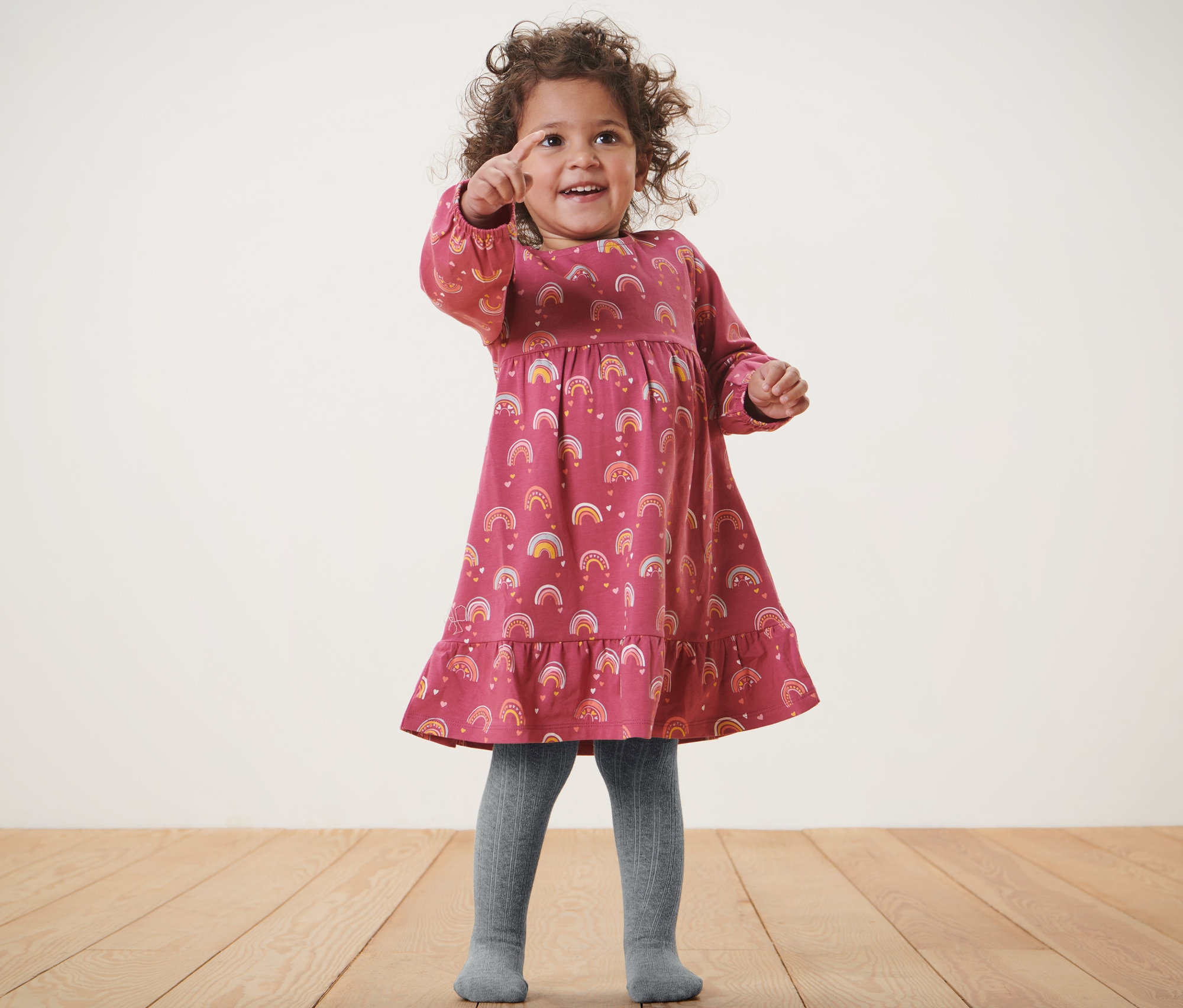 Dziewczynka w sukience w tęczowy wzór i szarych rajstopach niemowlęcych, 2 pary, pokazuje palcem.