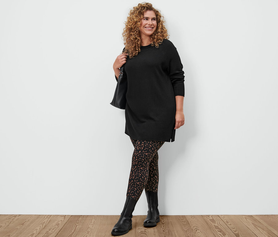 Kobieta pozuje w czarnym długim swetrze z dzianiny, legginsach z nadrukiem, butach skórzanych i z torbą na ramię.