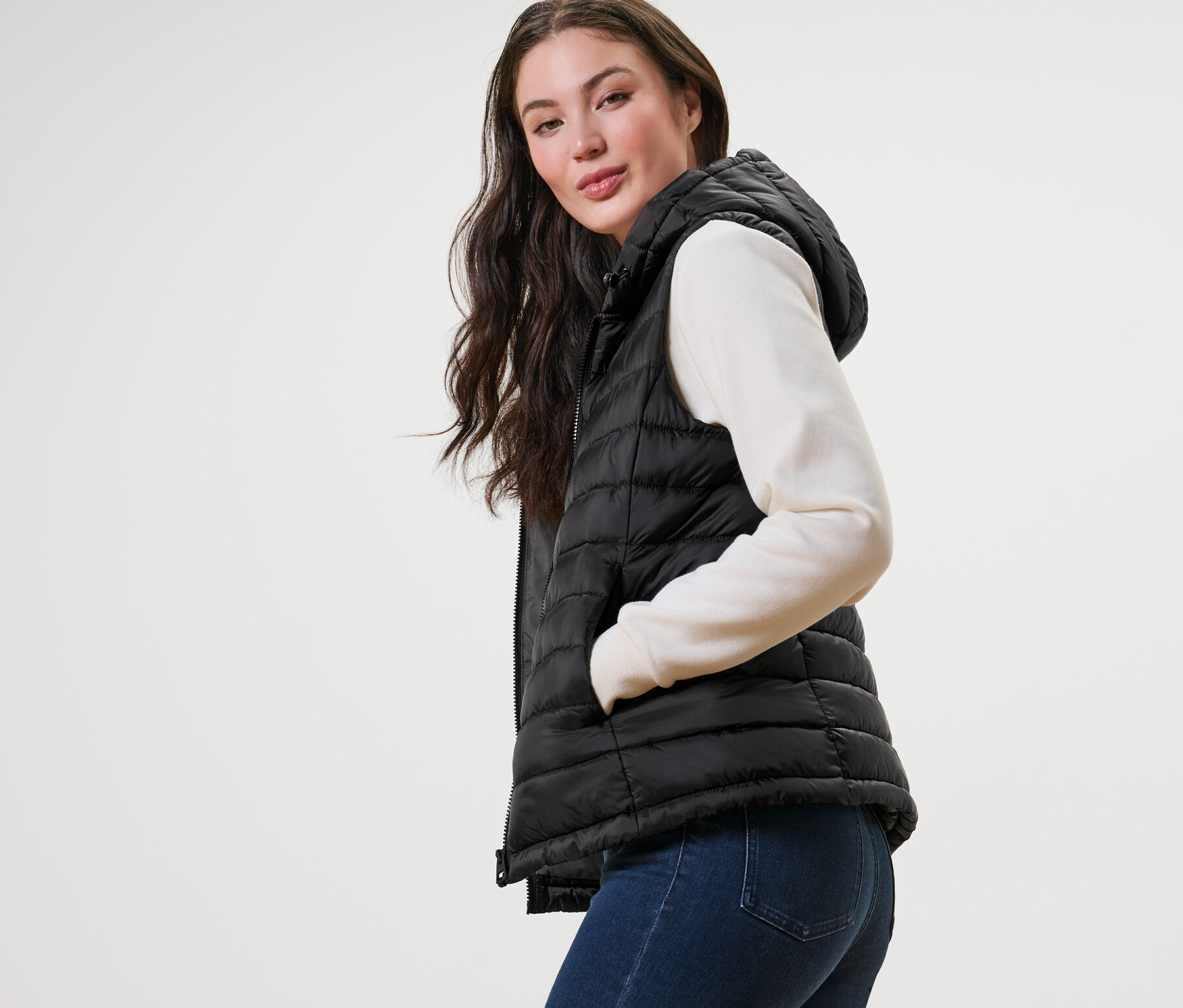 Kobieta pozuje w pikowanej kamizelce z kapturem, swetrze i dżinsach.