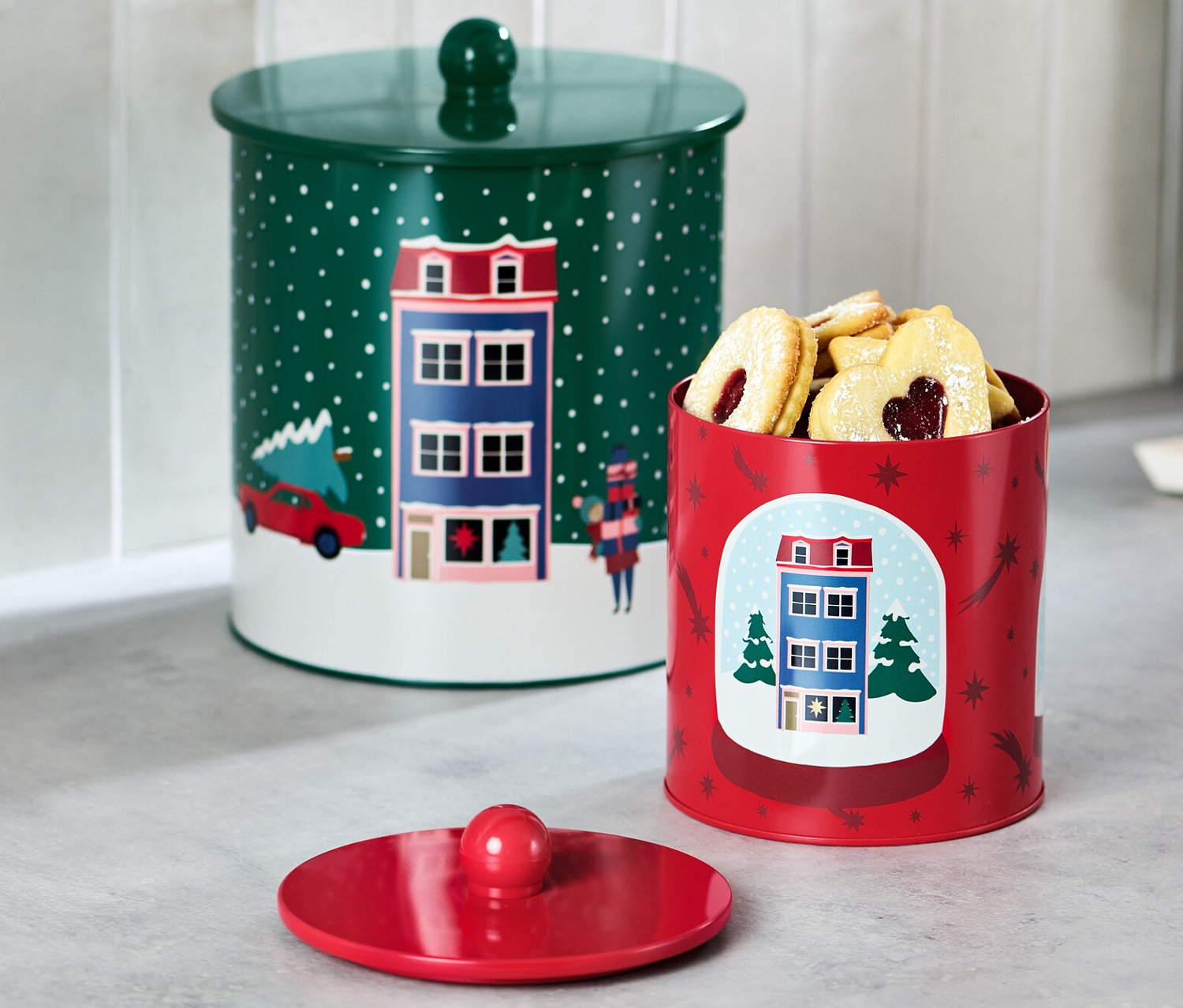 Na blacie kuchennym stoją dwie puszki na ciastka. Jedna puszka jest zielona z pokrywką, a druga czerwona i wypełniona ciasteczkami.