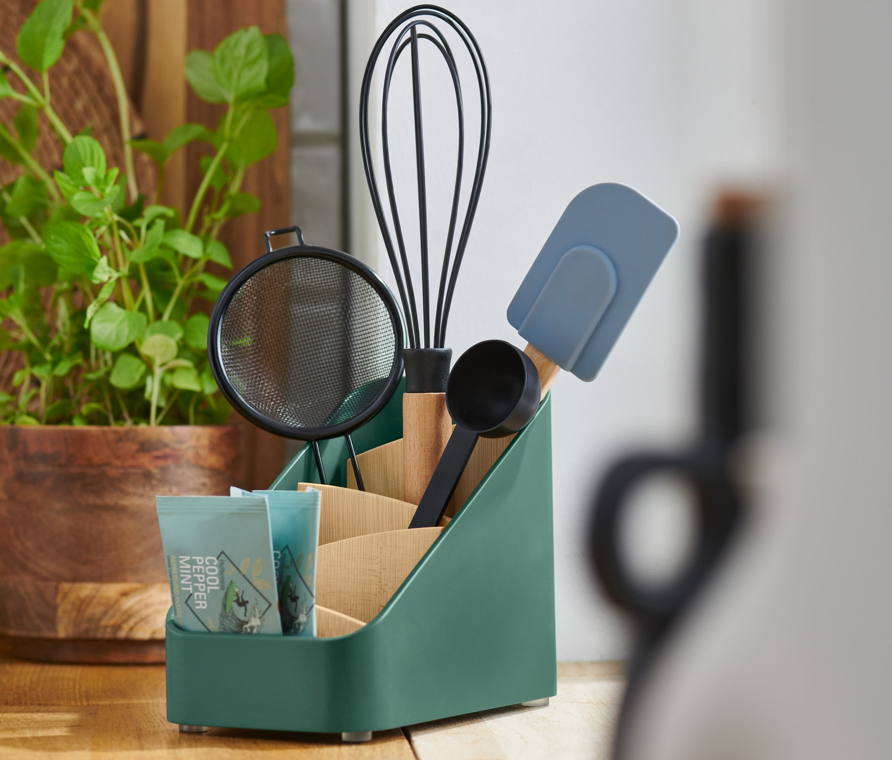 Zielony stojak na przybory kuchenne zawiera sitko, trzepaczkę, miarkę, szpatułkę i paczkę herbaty.