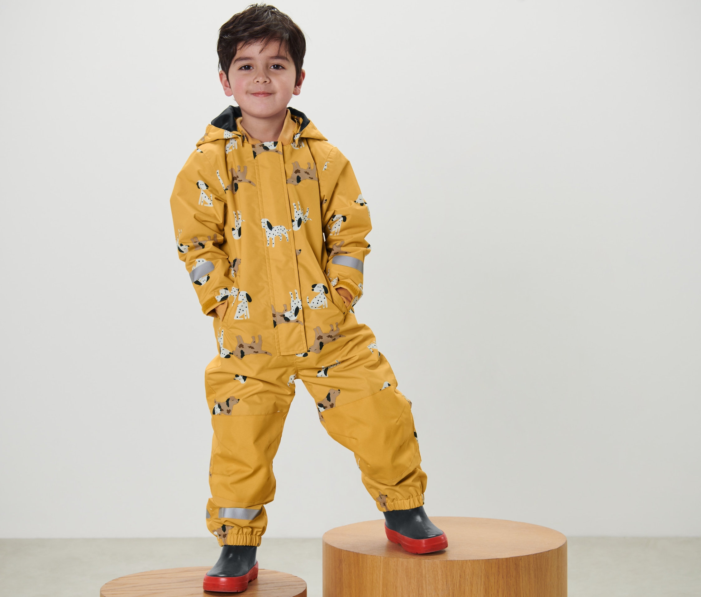 Chłopiec stoi na drewnianym postumencie i ma na sobie żółty kombinezon przeciwdeszczowy z nadrukiem psów.