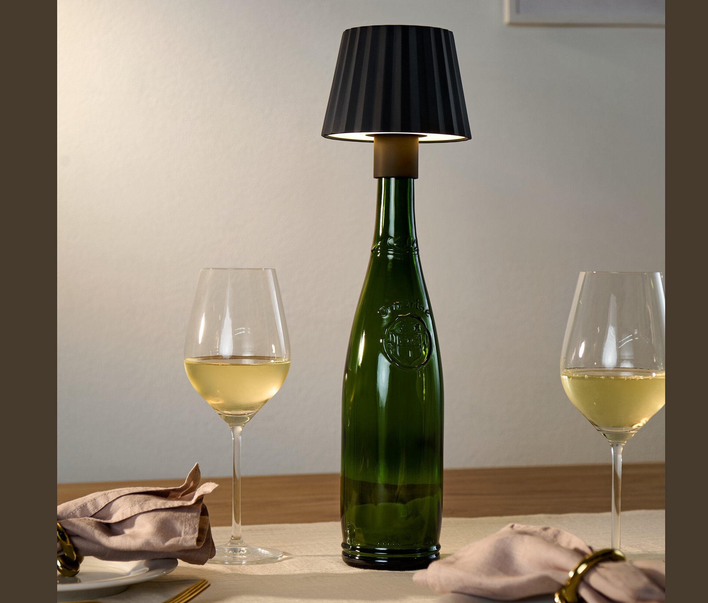 Na stole stoi zielona butelka z czarną lampą LED na butelkę, obok dwa kieliszki białego wina.