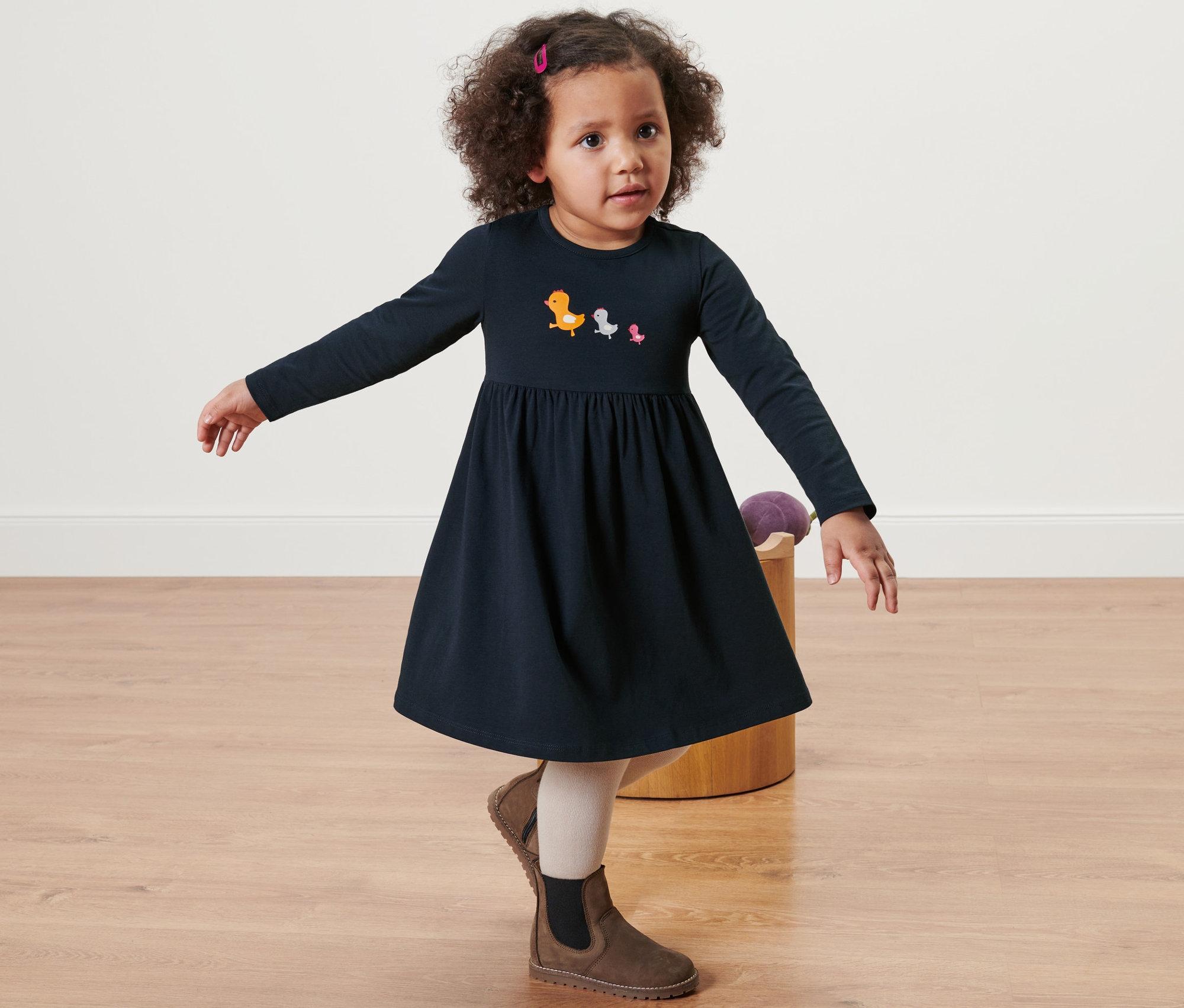 Dziewczynka pozuje w granatowej dżersejowej sukience z długim rękawem i wzorami ptaków, do tego brązowe buty.