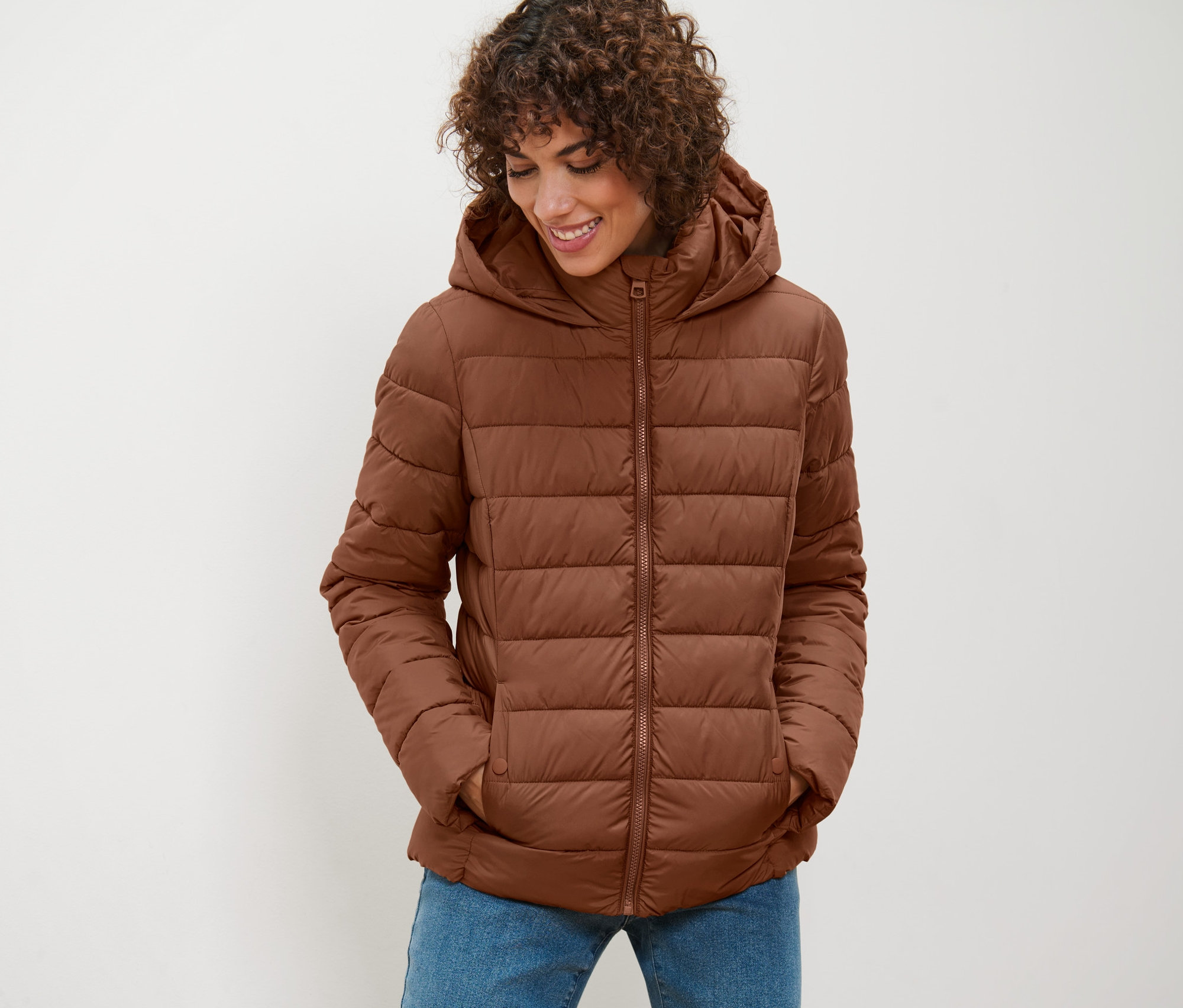 Kobieta ma na sobie koniakową kurtkę pikowaną z kapturem i patrzy w dół.