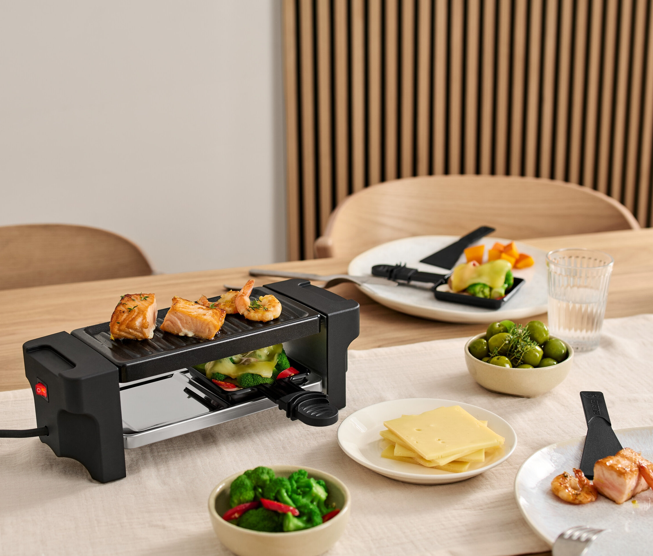 Raclette dla dwóch osób stoi na stole z łososiem i krewetkami na grillu. Talerze ze składnikami stoją wokół grilla.