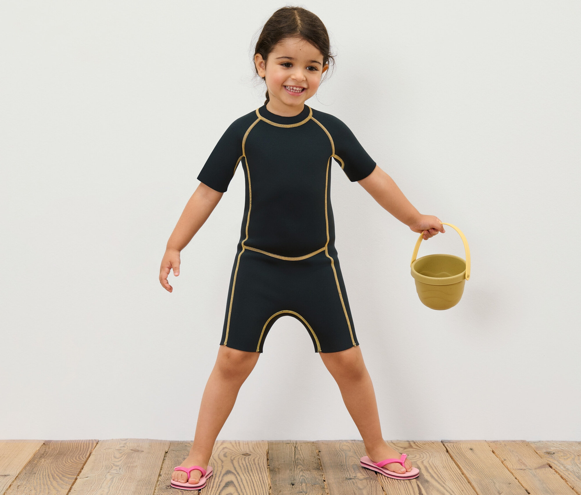 Dziewczynka w dziecięcym kombinezonie piankowym trzyma żółte wiaderko.