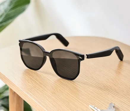 Czarne okulary przeciwsłoneczne ze słuchawkami Bluetooth® leżą na drewnianym stole obok kluczy i portfela.