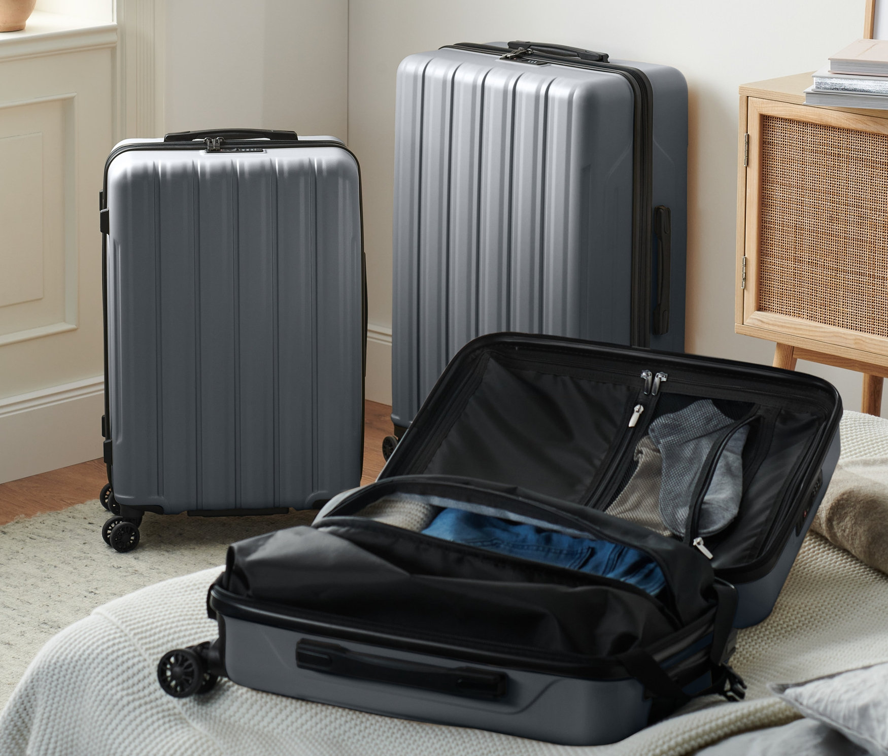 Trzy szare twarde walizki (mała, średnia i duża) stoją w pokoju, jedna otwarta na łóżku.