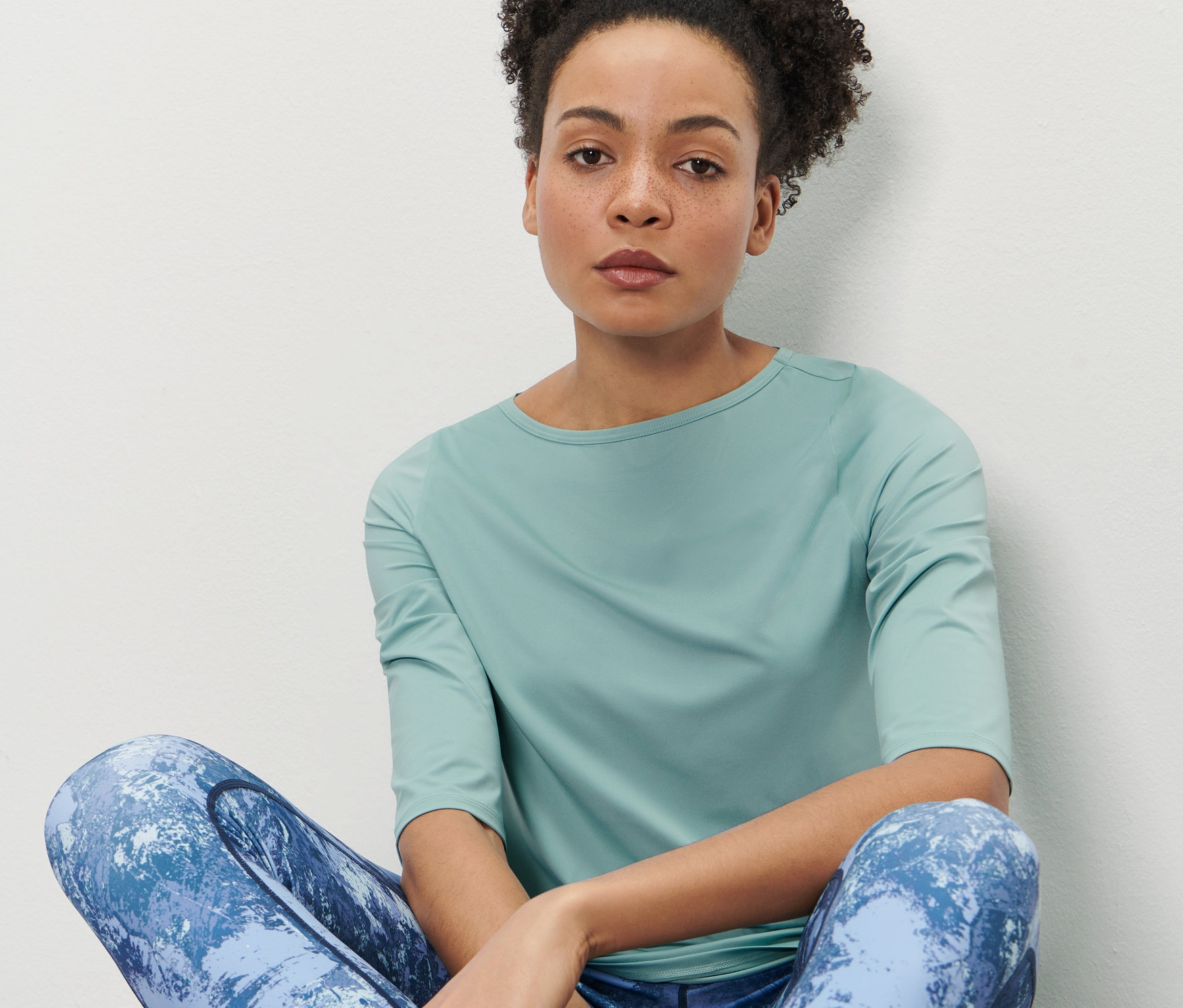 Kobieta siedzi i ma na sobie miętową koszulkę funkcyjną z rękawami o długości 3/4.