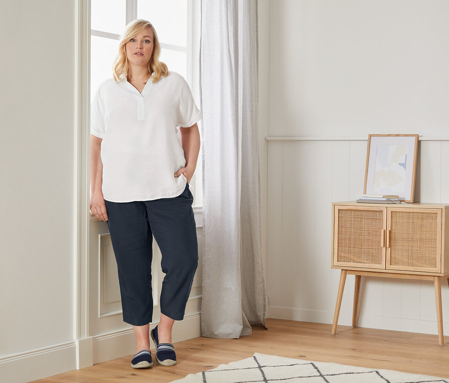 Kobieta pozuje przy oknie w bluzce oversized, luźnych spodniach chino i espadrylach.