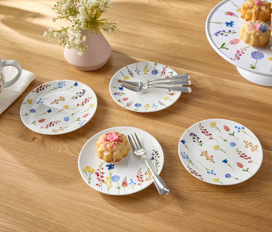 Na drewnianym stole stoi filiżanka kawy, wazon z kwiatami, talerze i małe ciastka.