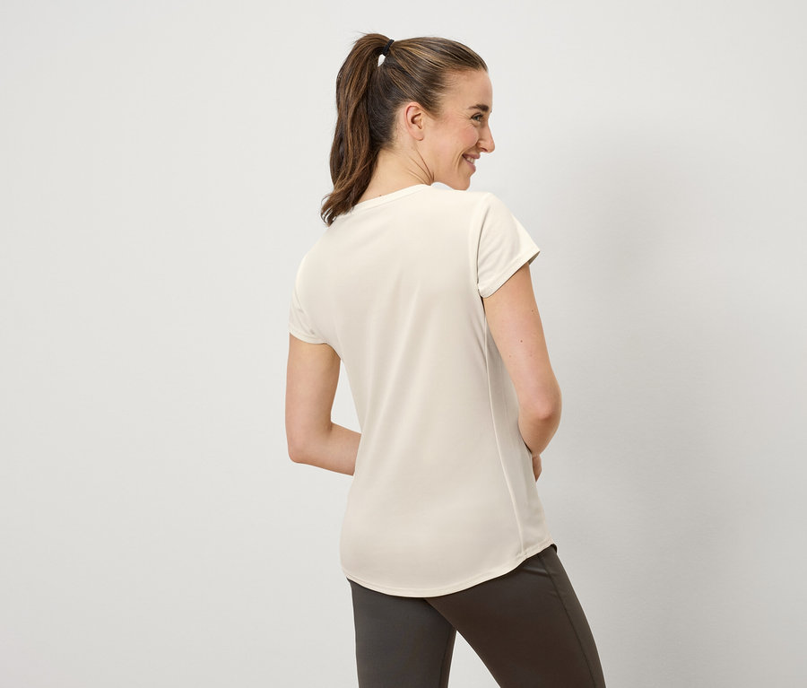 Kobieta z brązowymi włosami związanymi w kucyk uśmiecha się i patrzy w lewo. Ma na sobie kremową koszulkę i brązowe spodnie.