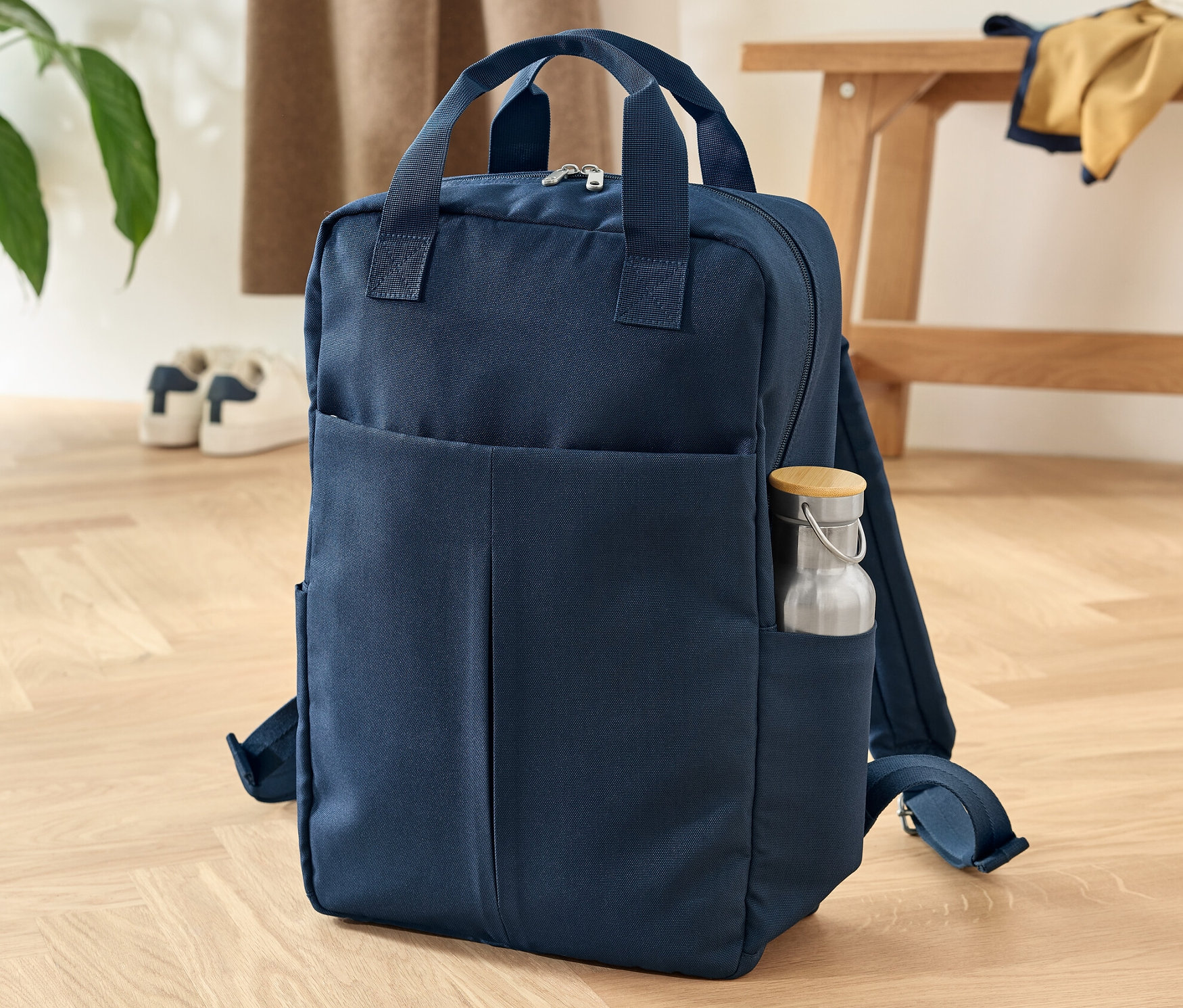 Granatowy plecak ze specjalnym zabezpieczeniem stoi na drewnianej podłodze. W bocznej kieszeni znajduje się butelka z wodą.