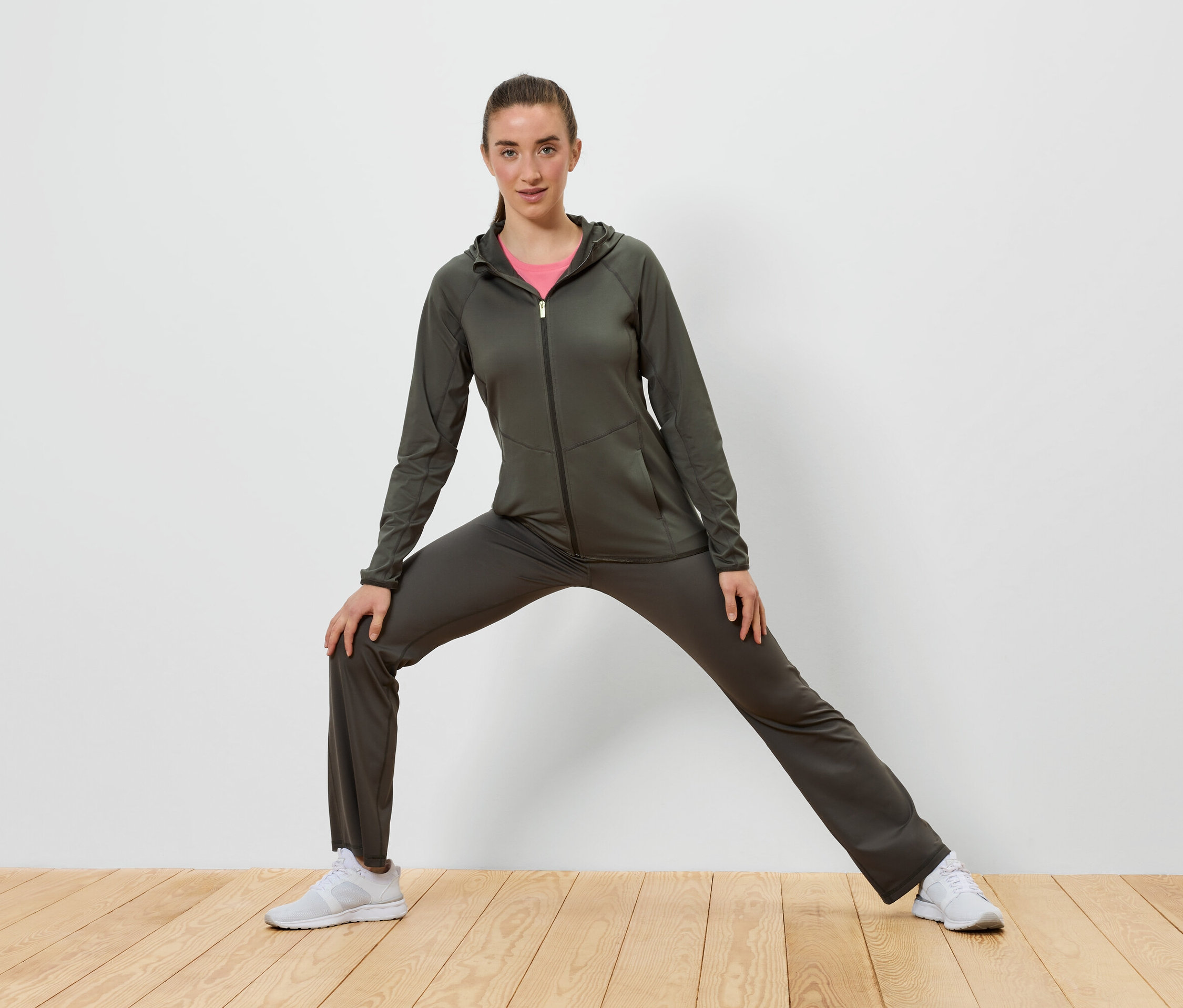 Kobieta w oliwkowym dresie rozciąga się na drewnianej podłodze przed białą ścianą.