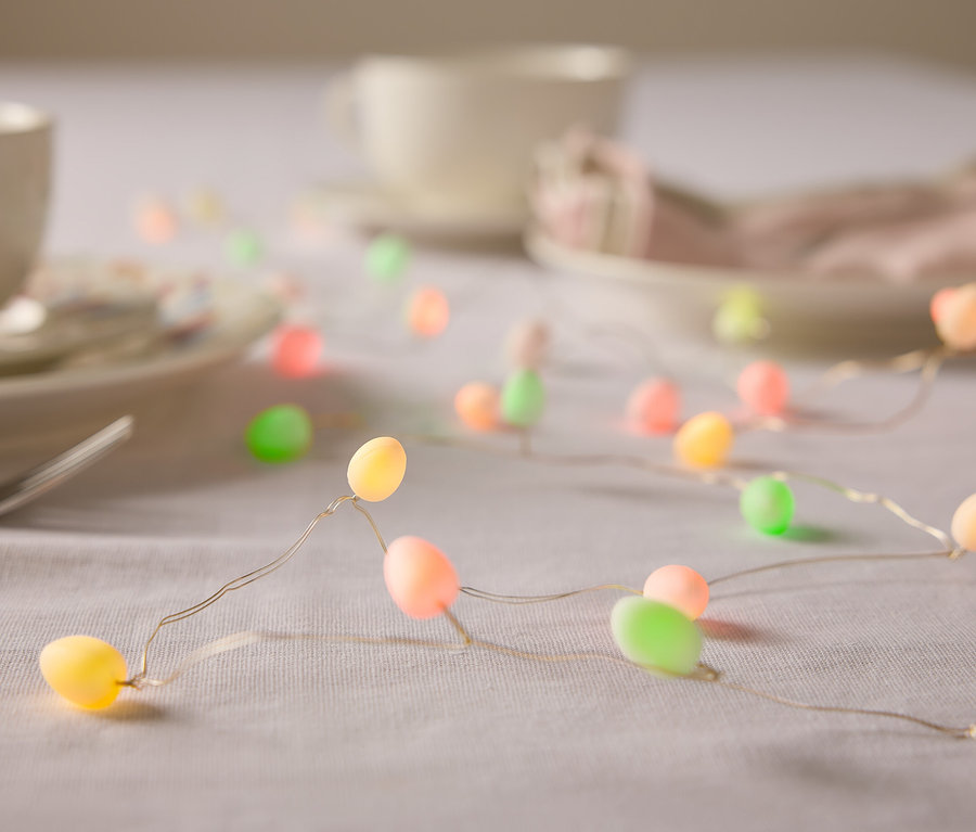 Zbliżenie stołu z naczyniami i łańcuchem światełek w kształcie jajek wielkanocnych w pastelowych kolorach.