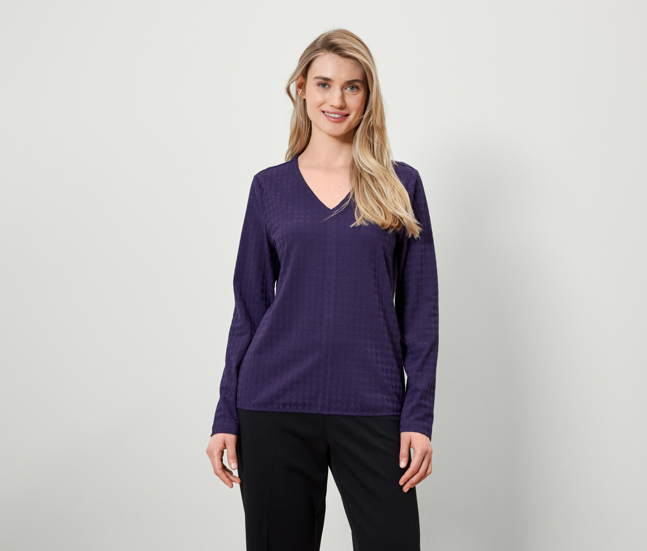 Blondynka z długimi włosami pozuje w fioletowej bluzce z długim rękawem i czarnych spodniach.