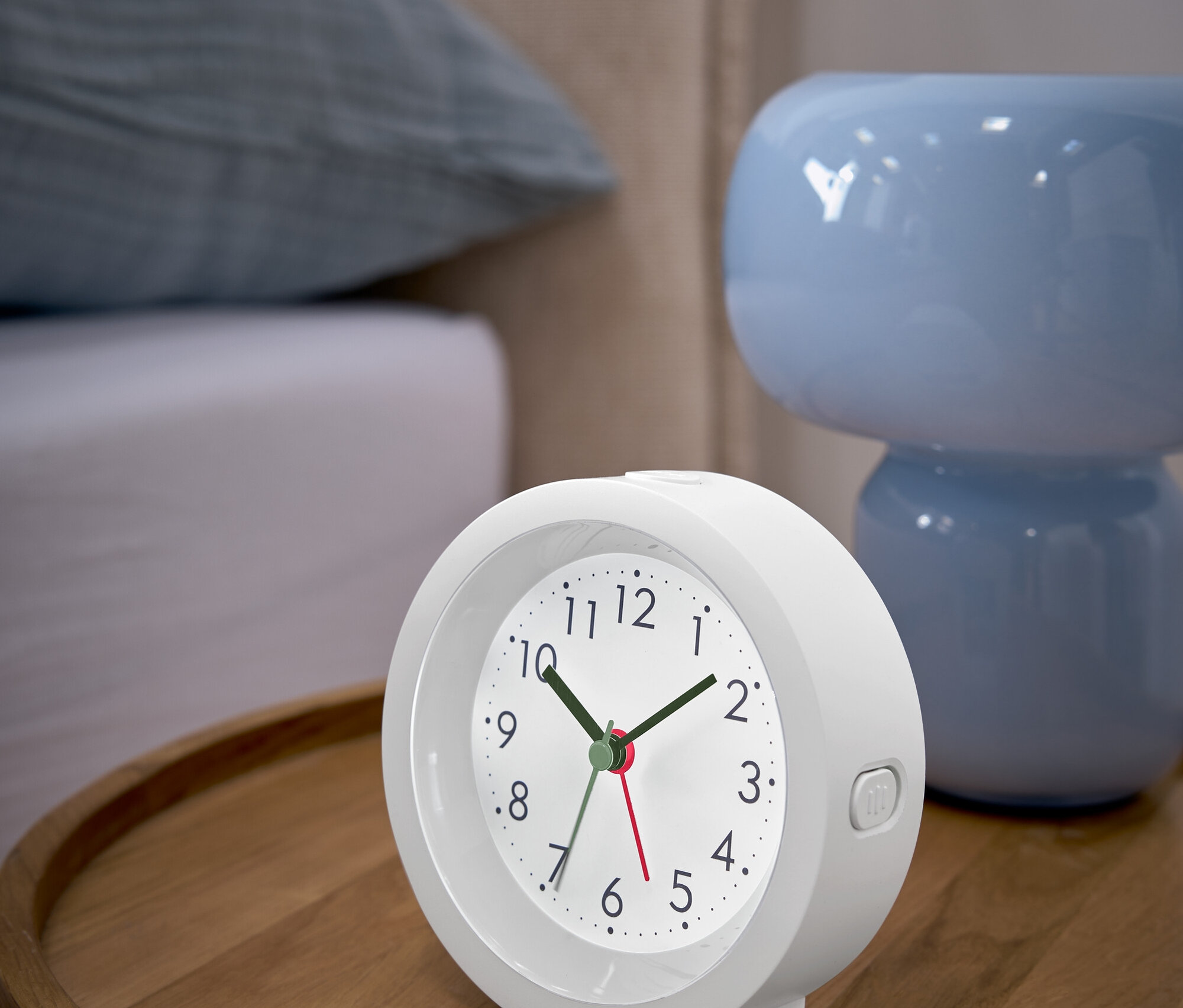 Biały budzik stoi na drewnianym stoliku nocnym obok niebieskiej lampki.