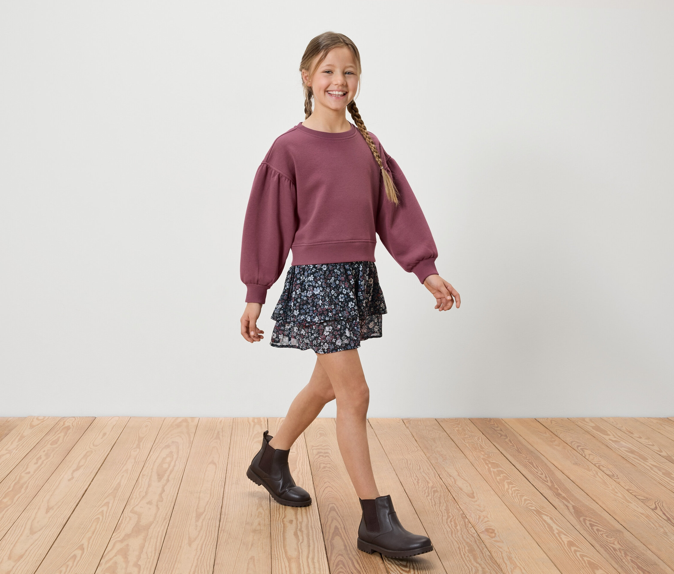 Uśmiechnięta dziewczyna z warkoczem idzie po drewnianej podłodze w bordowej bluzie, kwiecistej spódnicy i czarnych butach.