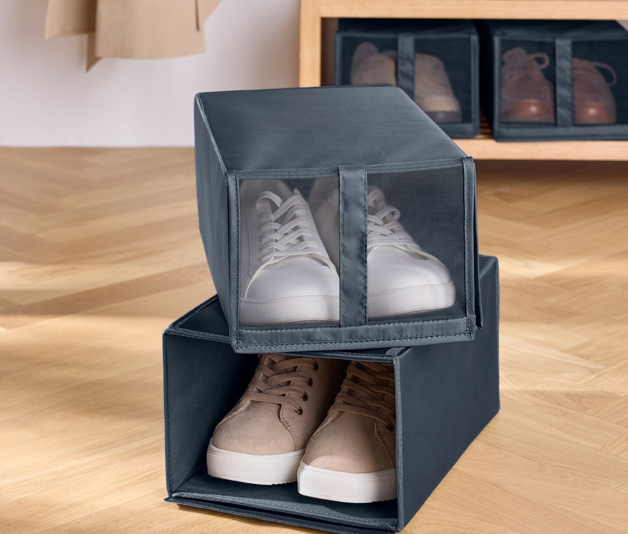 Dwa szare, materiałowe pudełka na buty ułożone jeden na drugim. W górnym pudełku znajdują się białe tenisówki, a w dolnym brązowe. W tle drewniana półka z butami.