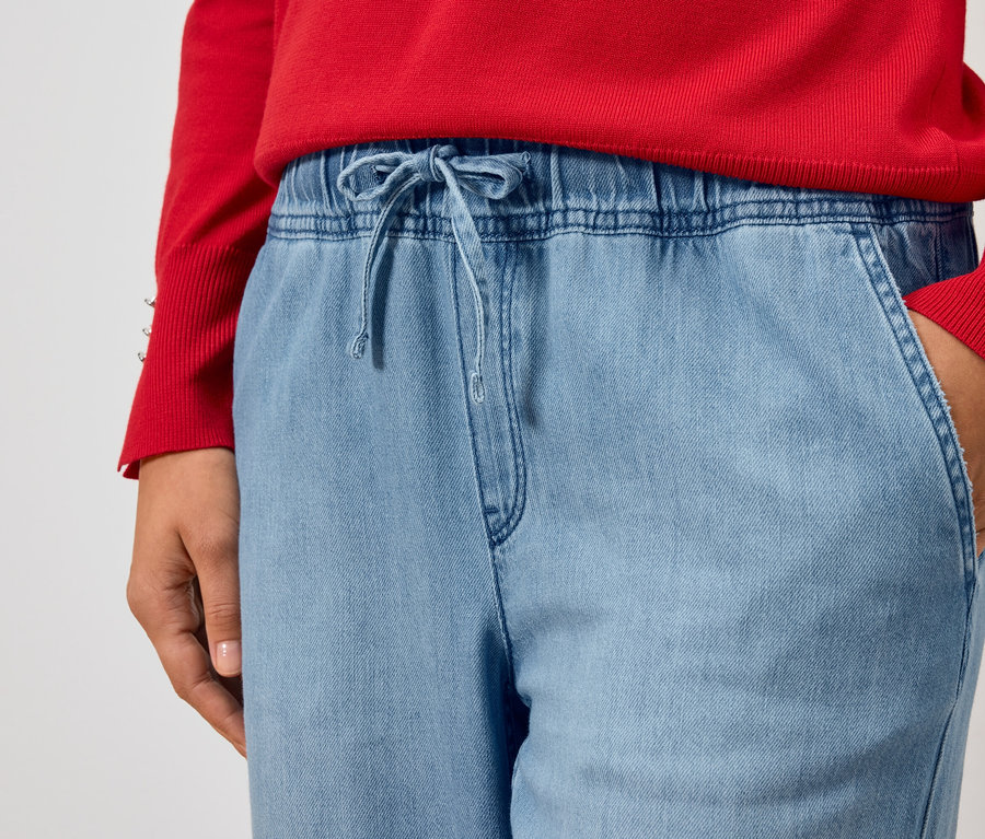 Częściowy widok osoby w czerwonym swetrze i jasno niebieskich dżinsach z ręką w kieszeni.