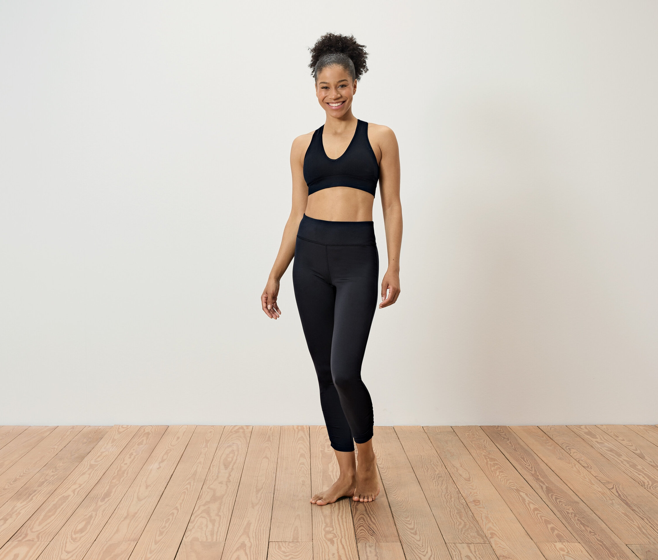 Kobieta w czarnym stroju sportowym stoi na drewnianej podłodze przed białą ścianą.